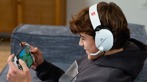 En person med et hvidt headset sidder i en sofa og er fuldt fokuseret på sin håndholdte spillekonsol, mens en automatisk klædefunktion gemmer deres spilfremskridt i baggrunden.