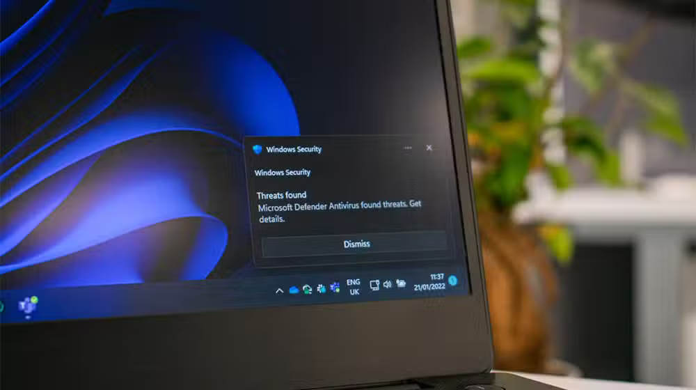 På skærmen på en bærbar computer vises en Windows-sikkerhedsadvarsel med teksten »Microsoft Defender Antivirus har fundet trusler«, med et blåt, abstrakt baggrundsbillede og nogle planter, der er slørede i baggrunden.